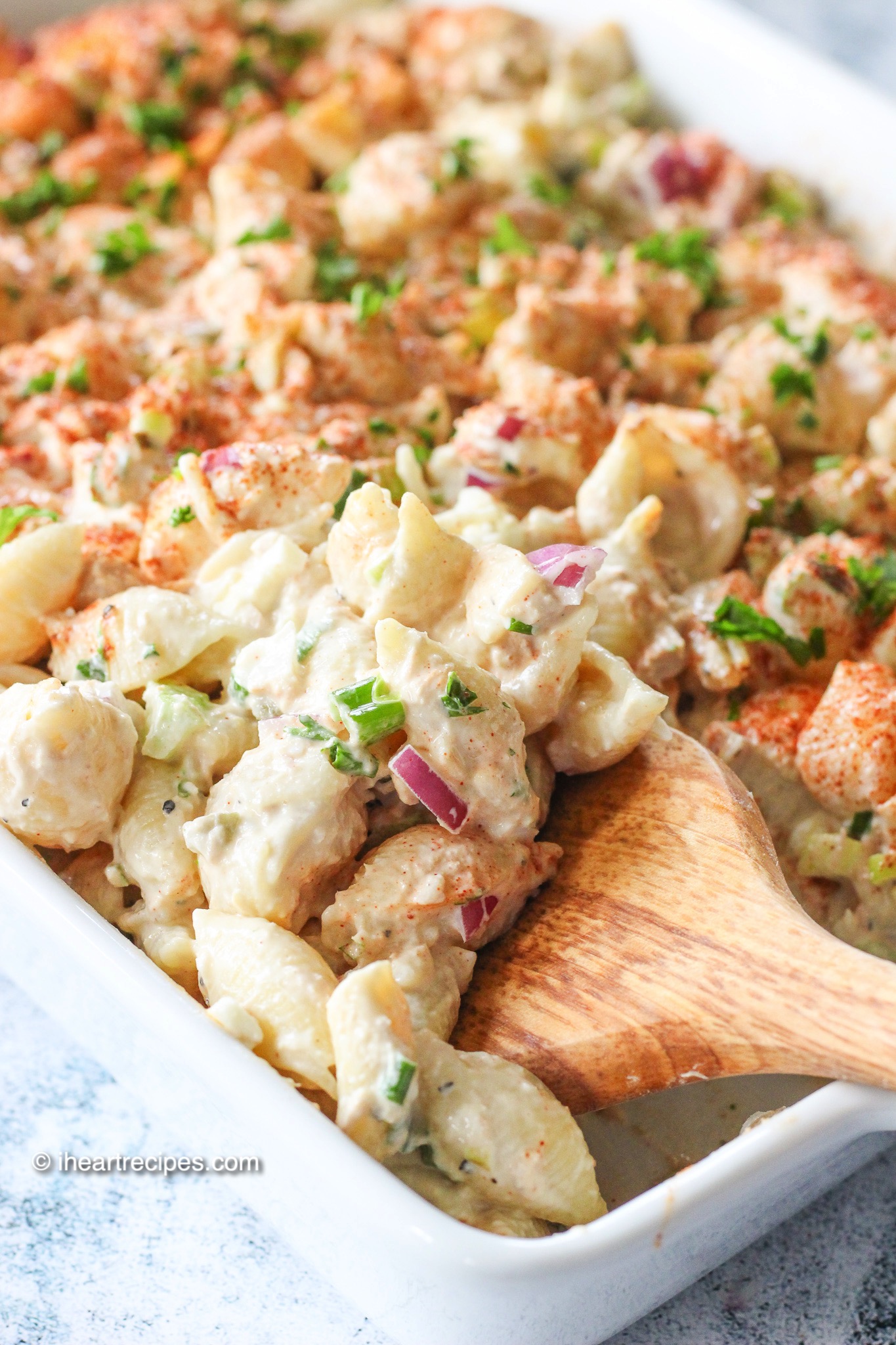 A close-up image of tuna pasta salad in a dish garnished with paprika, fresh parsley flakes and served with a wooden spoon.