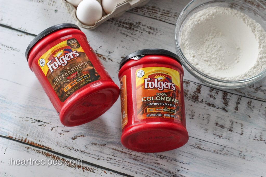 Two red containers of Folgers coffee  alongside a bowl of flour and a carton of eggs on a distressed white wooden table. 