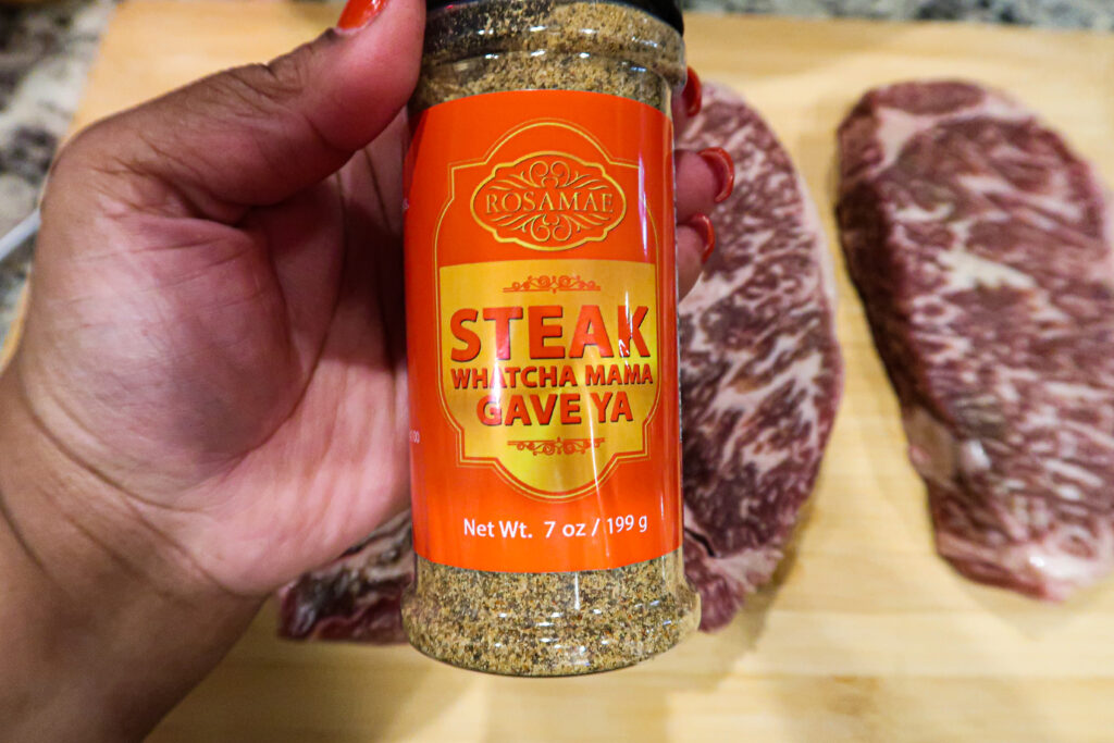 A close-up of a bottle of Rosamae "Steak Whatcha Mama Gave Ya" Seasoning. Rows of wagyu steaks sit on a wooden board in the background 