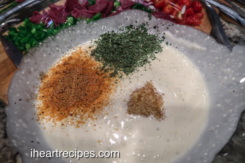 A large glass bowl holds a creamy Italian salad dressing mixture seasoned with seasoning salt, basil, and minced garlic.