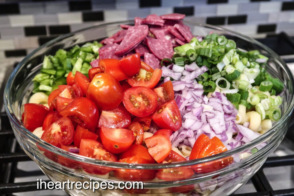 All the fresh ingredients needed to make an Italian pasta salad in a large glass bowl. Chopped tomatoes, red onions, green onions, peppers, and sliced salami fill the bowl.