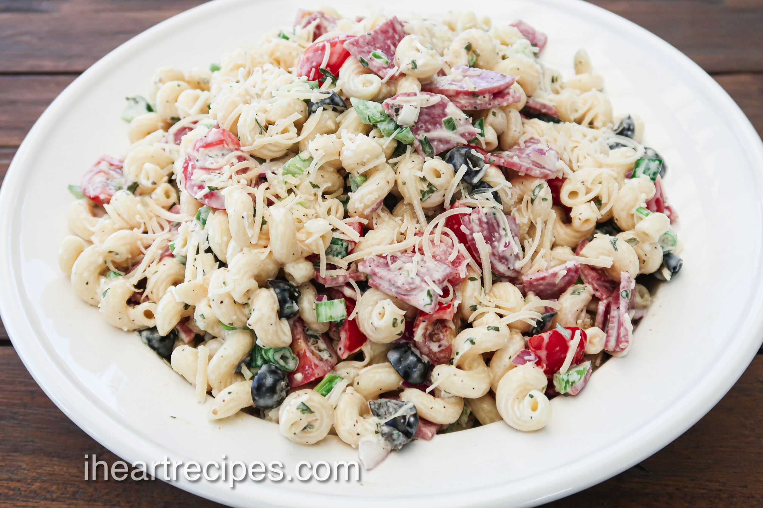 A large white bowl filled with Italian pasta salad—corkscrew pasta mixed with a creamy dressing, shredded cheese, chunks of salami, and colorful vegetables.