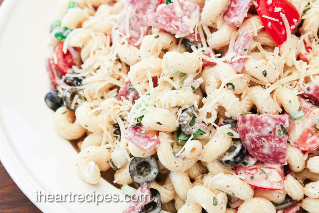 A close-up image of Italian pasta salad in a large white bowl. The pasta salad is made with cooked corkscrew pasta mixed with shredded cheese, olives, peppers, salami, herbs, and a creamy dressing.