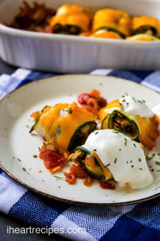 Three zucchini chicken taco rolls topped with sour cream and melted cheese on a bed of bright red salsa. A white casserole dish filled with rolls sits nearby. 