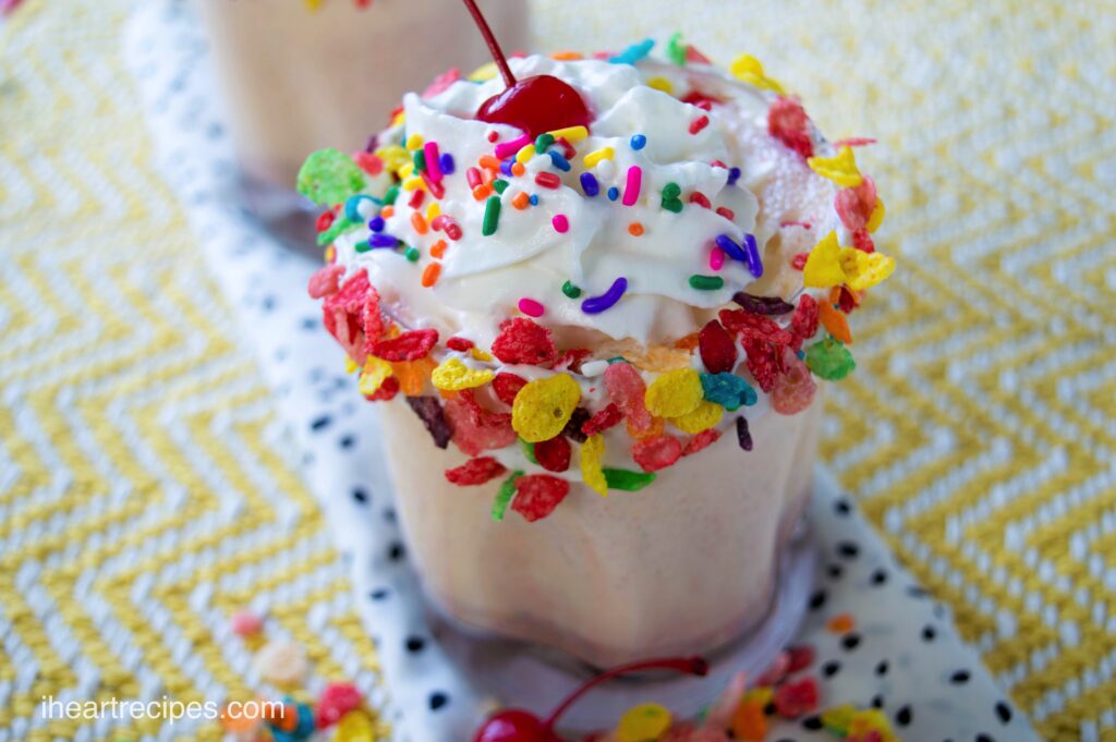 A sweet Fruity Pebbles milkshake in a glass, topped with whipped cream, sprinkles, Fruity Pebbles cereal, and a cherry.