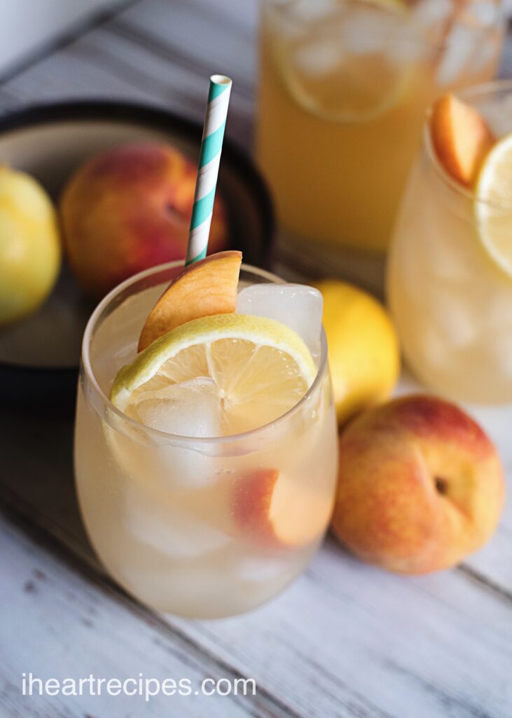 Refreshing glasses of ice-cold peach lemonade, garnished with fresh peach and lemon slices, surrounded by whole fresh peaches.