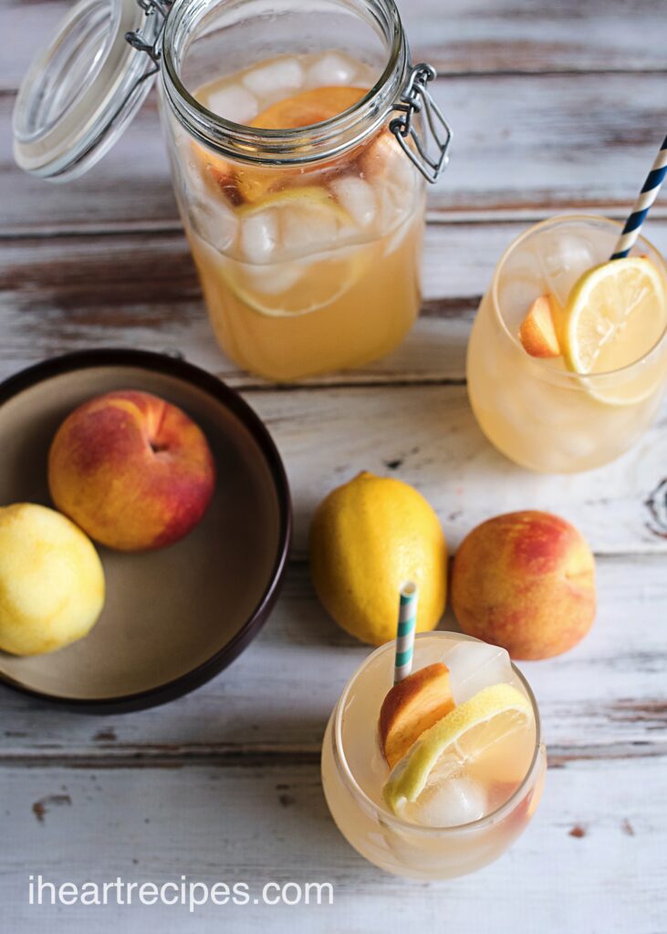 Glasses of fresh peach lemonade garnished with peaches and lemon slices, on a table with a bowl of fresh peaches.