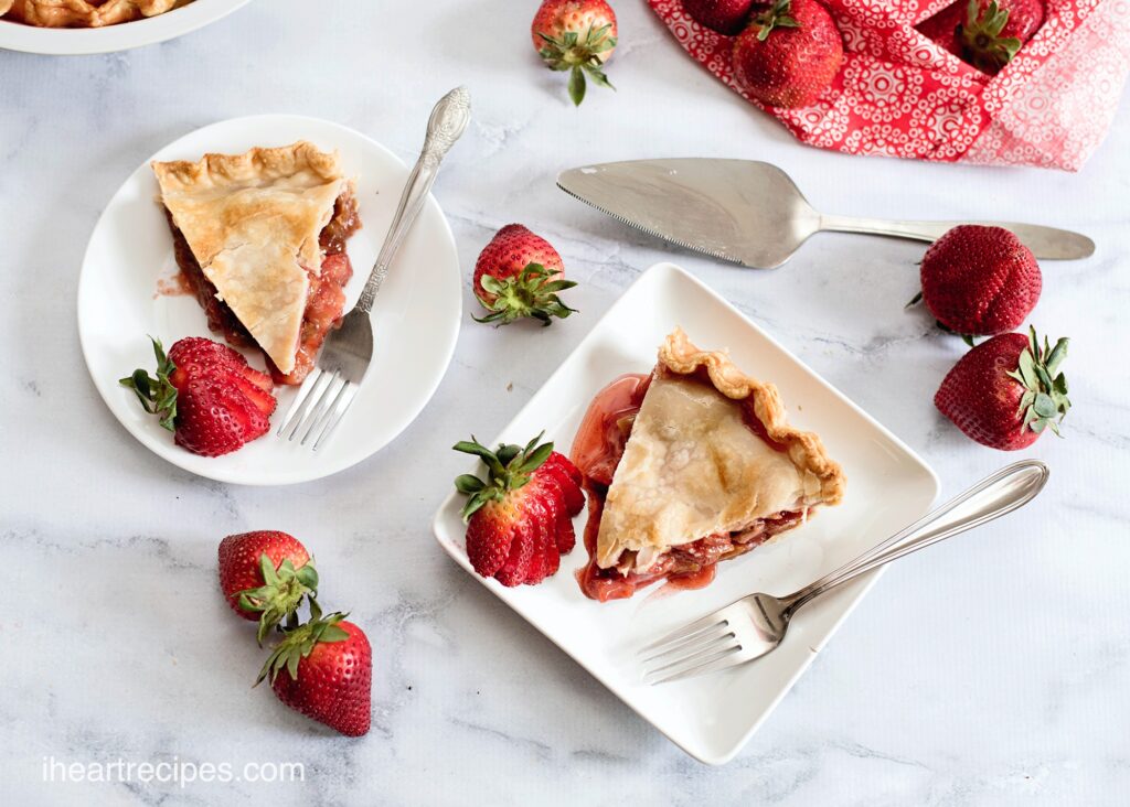 Two slices of fresh homemade strawberry rhubarb pie on white plates, surrounded by fresh strawberries and silverware.