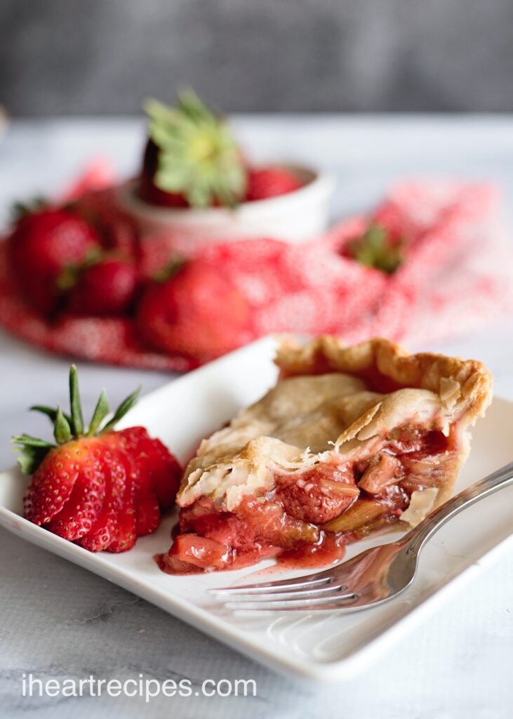 Side view of a slice of fresh-baked strawberry rhubarb pie served on a white plate with a fresh sliced strawberry.