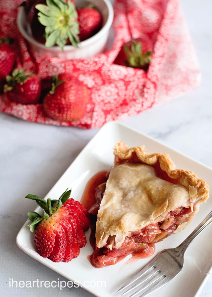 A single slice of homemade strawberry rhubarb pie served on a while plate with a fresh strawberry garnish.