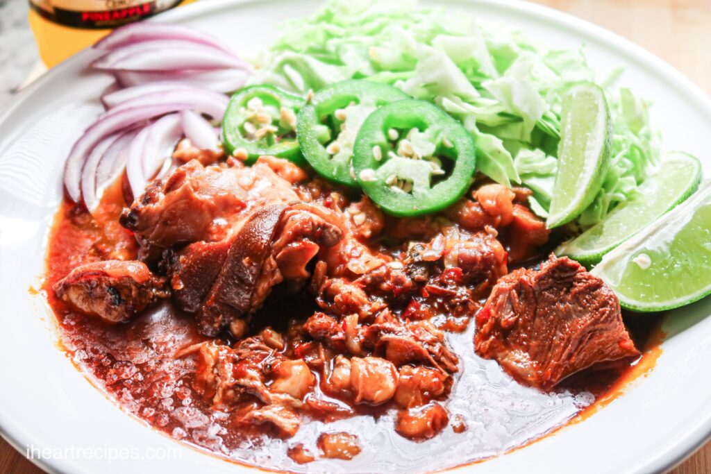 A flavorful pork pozole stew served with lime, jalapeno, and red onion slices and fresh cabbage assembled in a white bowl.