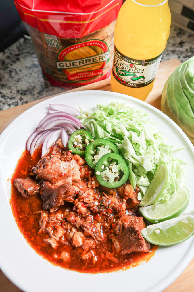 Flavorful pork pozole in a rich broth garnished with onions, jalapenos and limes arranged in a white bowl. A pineapple drink and tostadas sit nearby.  