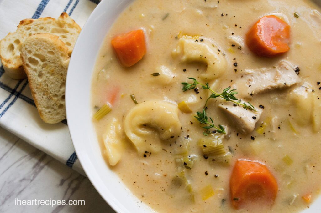 A bowl of creamy Chicken Tortellini soup served in a white bowl with sliced of toasted baguette.