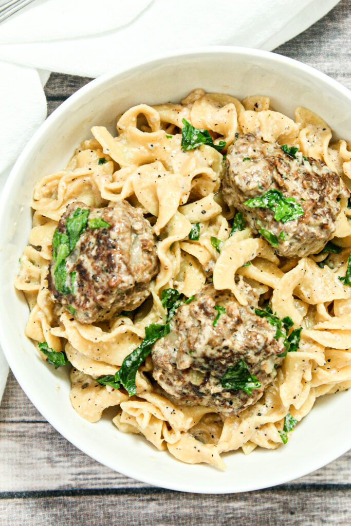 A white bowl filled with creamy egg noodles and three Swedish meatballs, topped with fresh parsley and grated parmesan cheese.