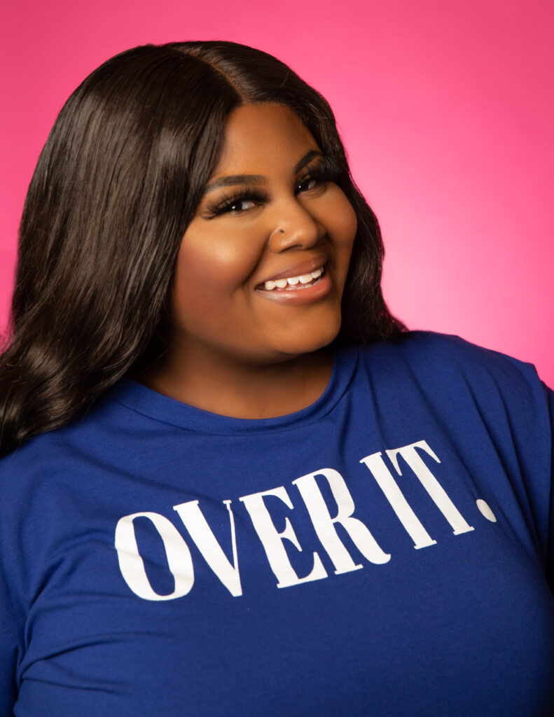 Rosie Mayes - an African American woman - is the author and voice behind I Heart Recipes. She's wearing a blue t-shirt with the words "over it" written on the front. She has long brown hair and is smiling at the camera.