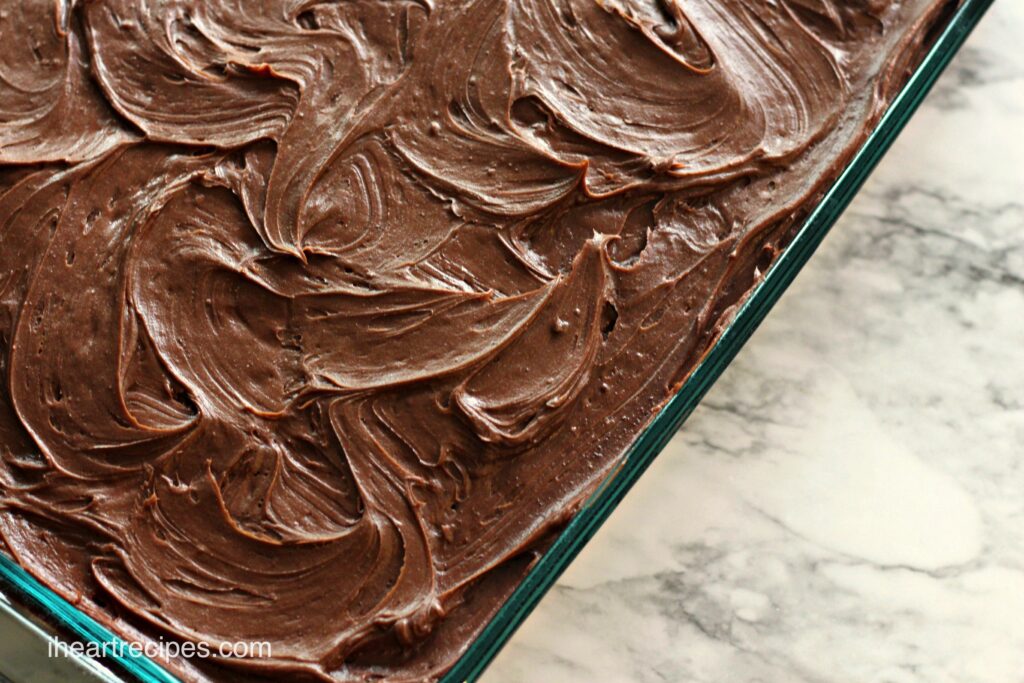 An overhead image of swirls of chocolate frosting on top of sour cream chocolate cake in a glass baking dish on a marble countertop.