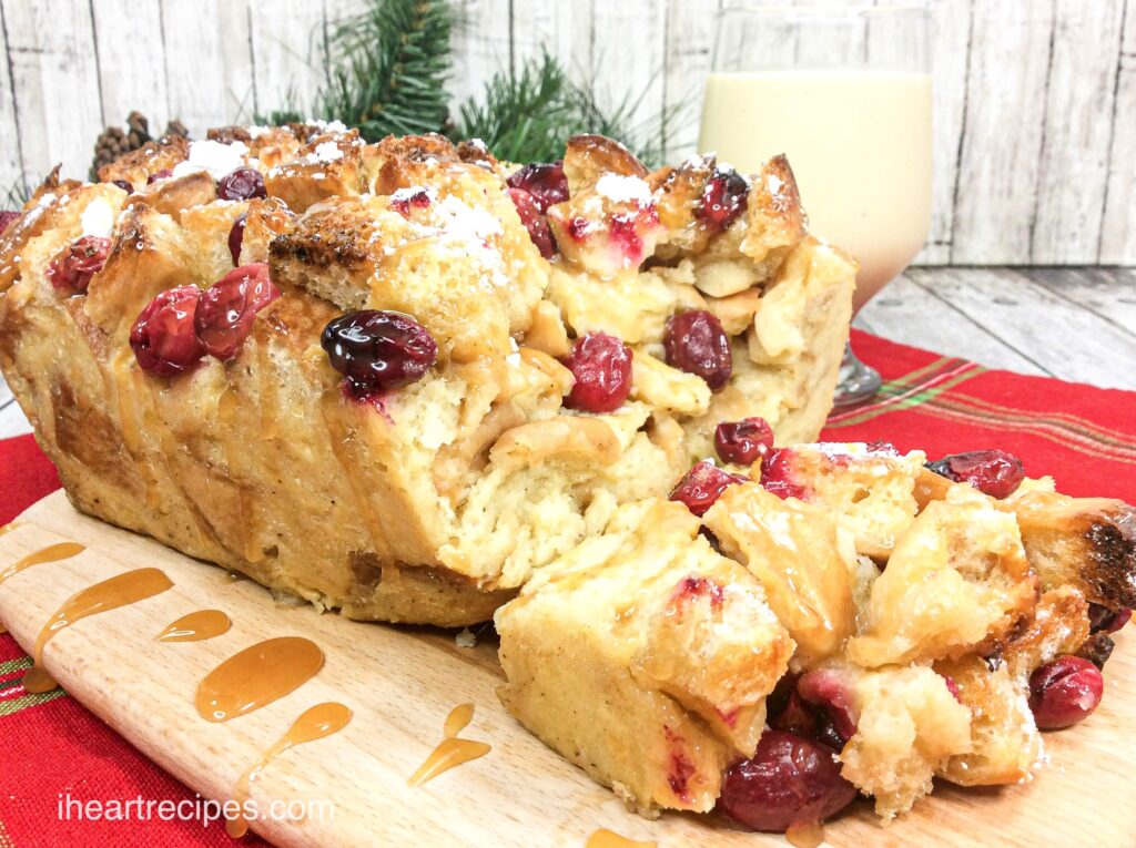 A loaf of eggnog bread pudding with a slice cut off, dusted with powdered sugar and packed with juicy cranberries.