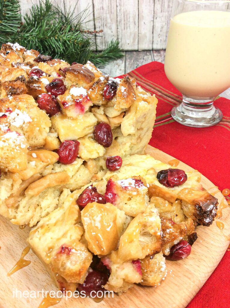 A close up look at a loaf of eggnog bread pudding, filled with juicy cranberries and topped with dusted powdered sugar, paired with a glass of creamy eggnog.