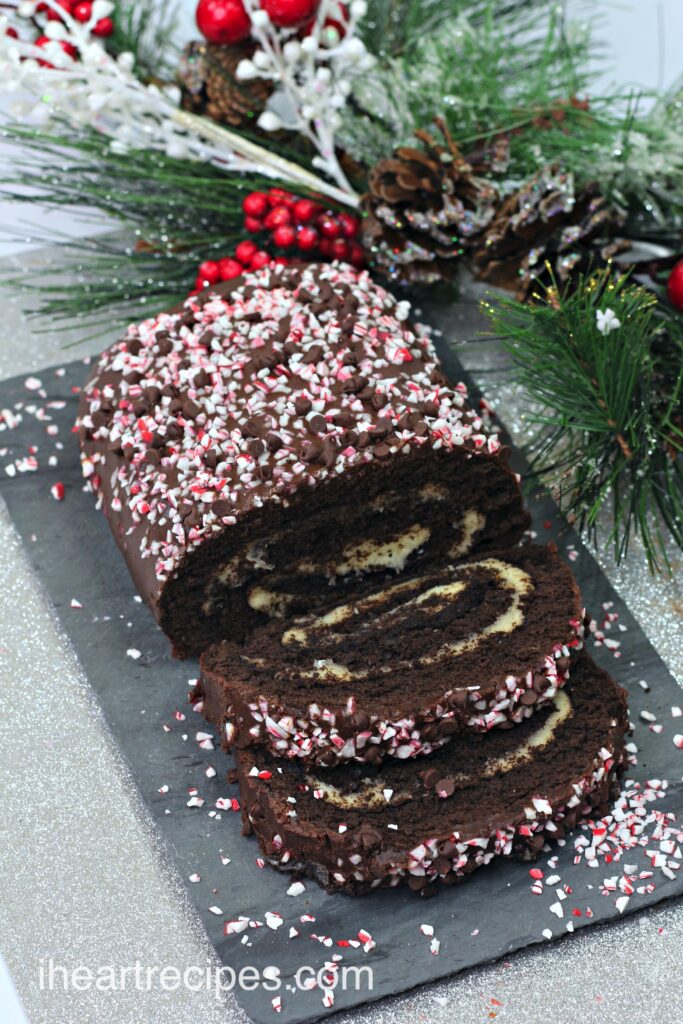 A chocolate peppermint Swiss rolls cake with two slices cut from the end, laying flat on a gray serving tray. The Swiss roll is filled with buttercream and topped with a chocolate ganache and crushed peppermint candy pieces.