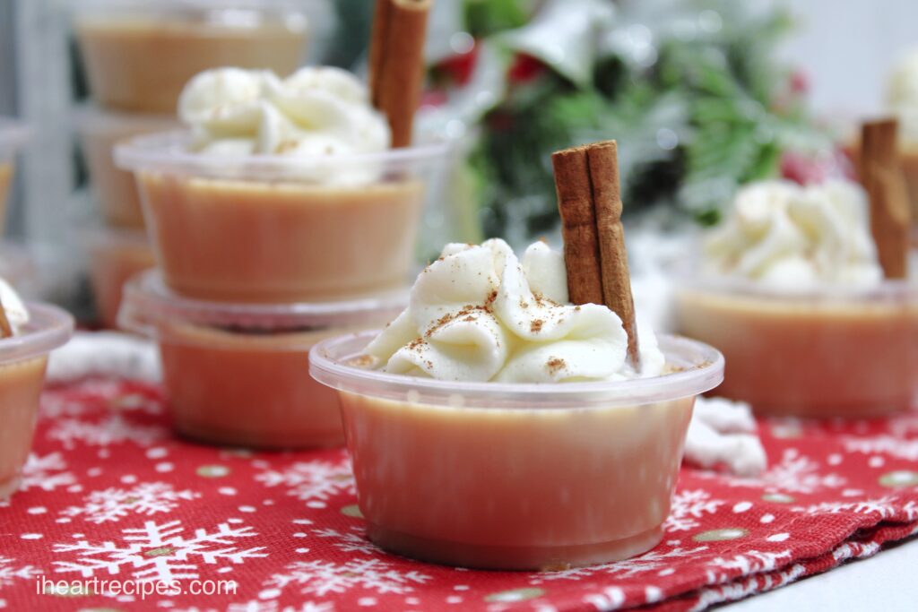 Small cups of eggnog jello shots topped with a dollop of whipped cream and a small cinnamon stick.