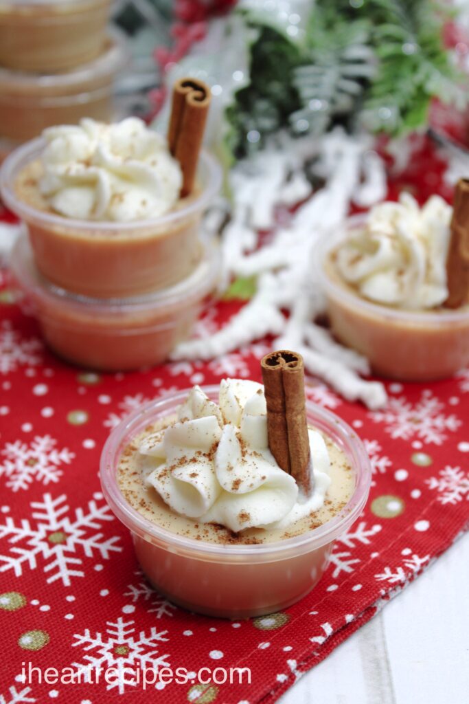 Eggnog Jello shots are topped with a sprinkle of cinnamon, a dollop of whipped cream, and a small cinnamon stick.