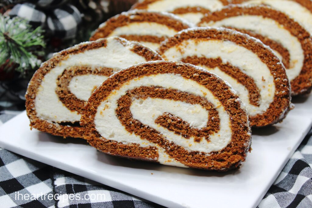 Slices of homemade gingerbread Swiss roll cake, filled with a homemade maple buttercream and topped with powdered sugar, served on a white plate.