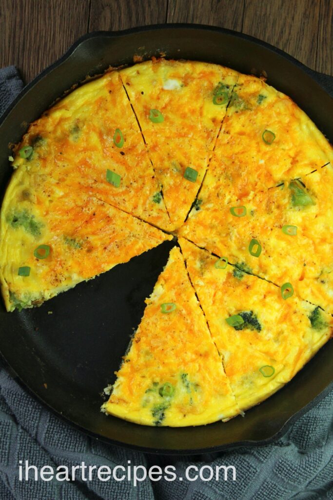 A large cast iron skillet holds a freshly baked breakfast frittata made with eggs, cheddar cheese, broccoli, and green onions. The frittata is sliced into 8 pieces, with one piece removed from the pan.