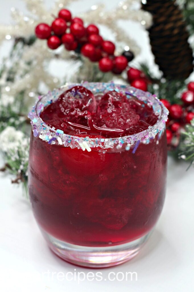 A Christmasy cocktail made with cherry vodka, gin, and fruit punch. The cocktail glass has a sugar and sprinkle rim and is garnished with a cherry.
