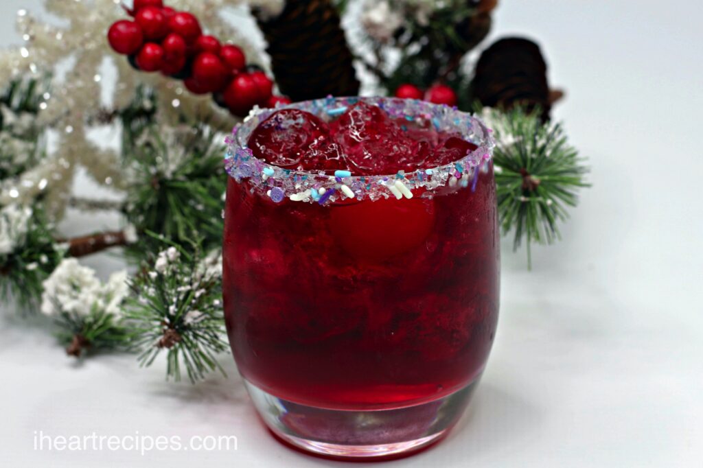 An easy Christmas cocktail in a glass. The cocktail is a deep Christmas red, with ice and a cherry, and a sugar sprinkle rim.