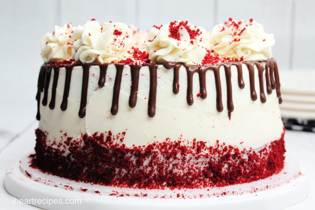 A whole red velvet cheesecake, iced with a cream cheese frosting and decorated with red velvet cake crumbles, chocolate ganache drizzle, and dollops of cream cheese frosting on top.