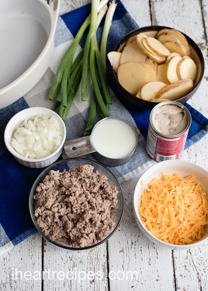 All the ingredients needed to make a hamburger and potato casserole, laid out in separate dishes. You'll need sliced potatoes, green onions, cream of mushroom soup, shredded cheese, cooked ground hamburger, onions and milk.
