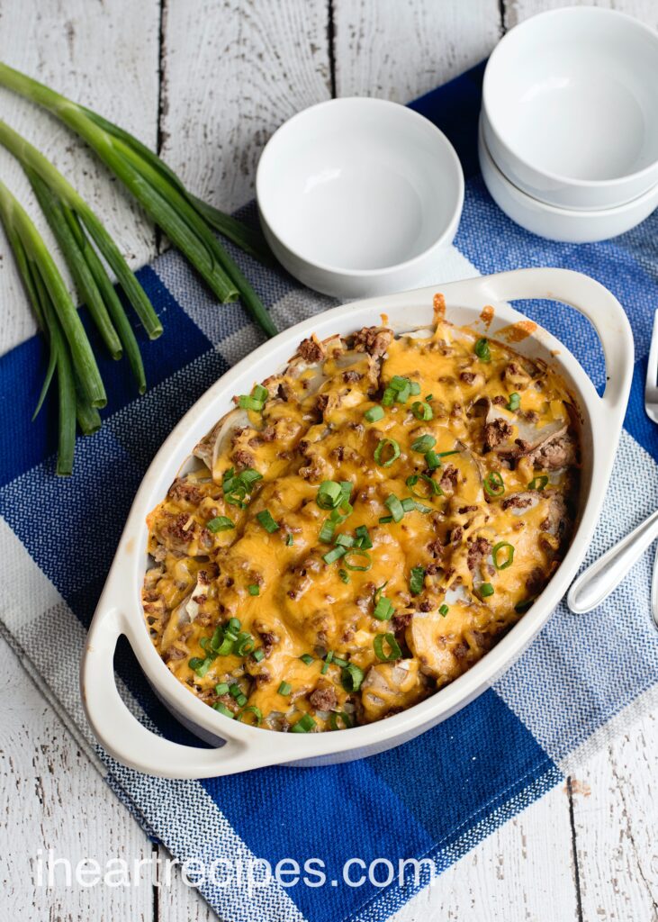 A white casserole dish filled with fresh-baked hamburger and potato casserole, with white bowls and stalks of green onions.