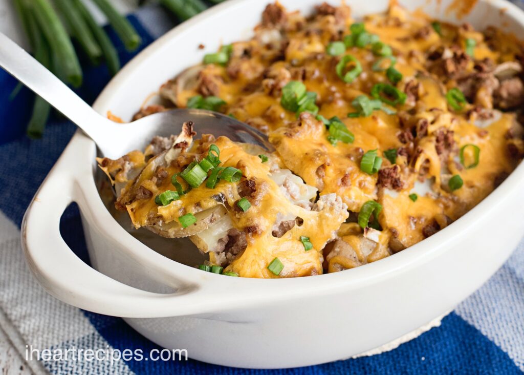 A hamburger and potato casserole, topped with cheese and green onions, in a white casserole dish. A silver serving spoon scoops out a single serving.