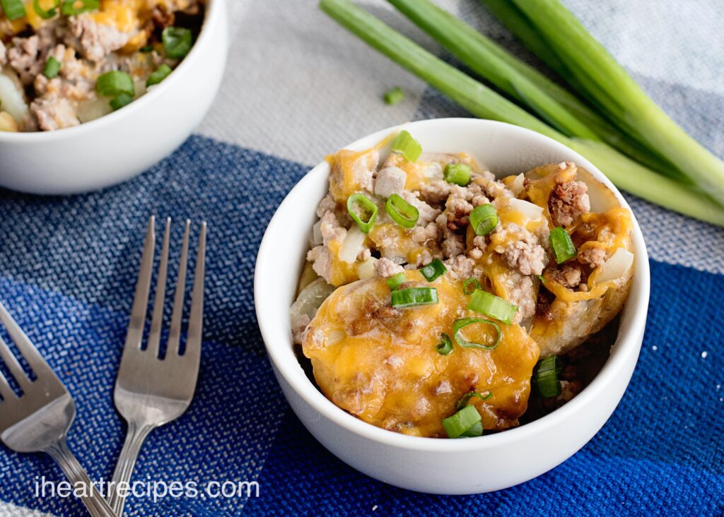 A white bowl full of hamburger and potato casserole, topped with cheddar cheese and green onions.