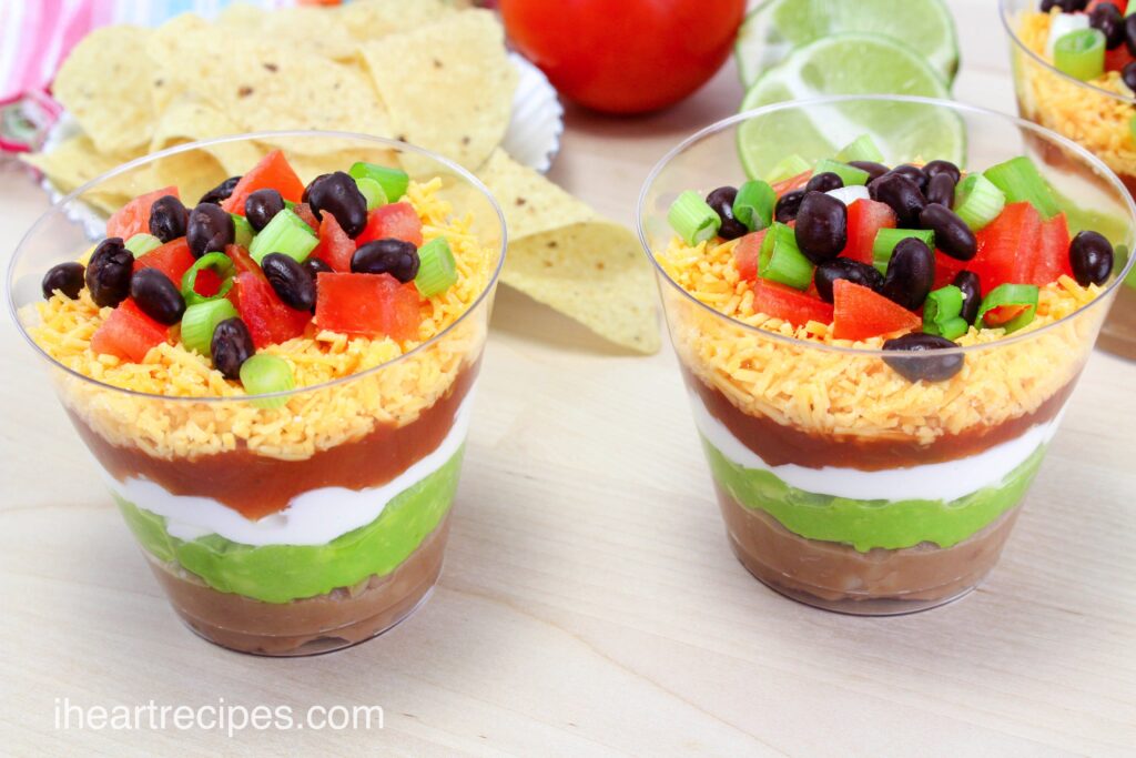 Individual nacho layer dip cups are layered with beans, guacamole, sour cream, salsa, cheese, and a tomato topping.
