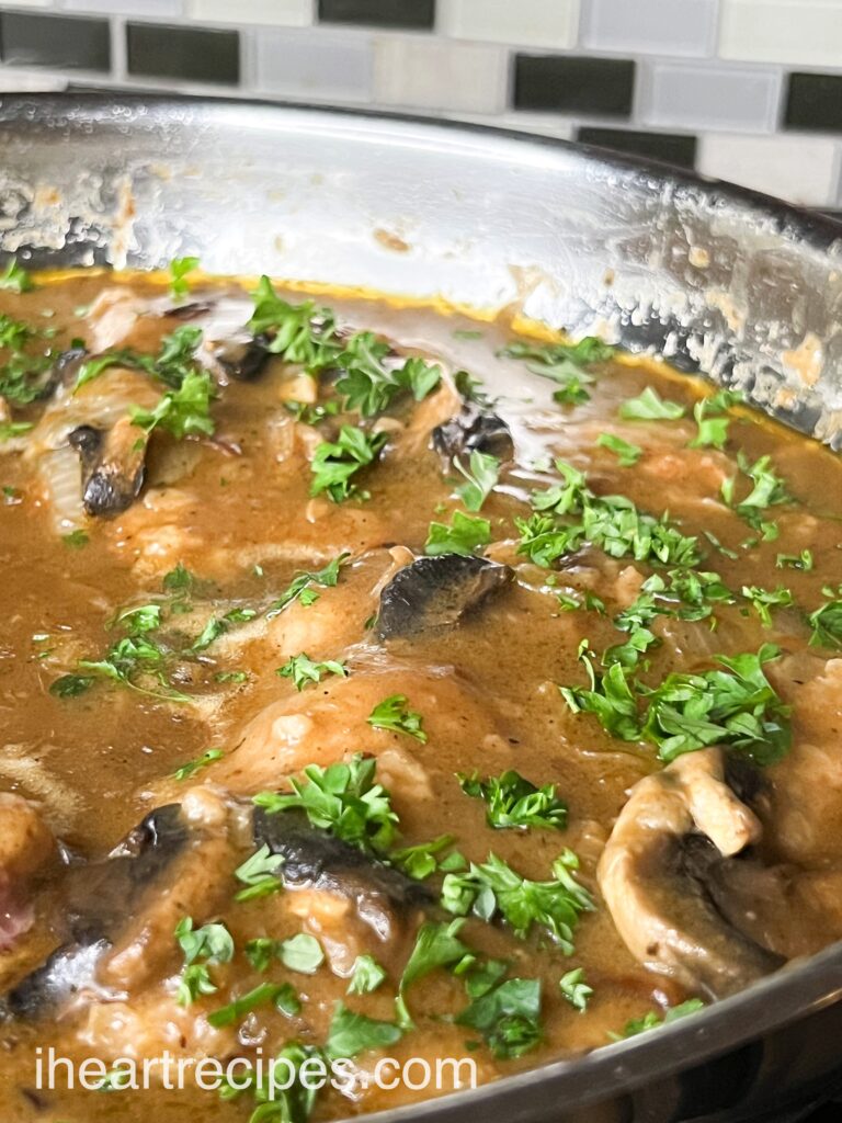 A close up image of a skillet filled with smothered pork chops in a brown mushroom gravy, garnished with fresh parsley leaves.