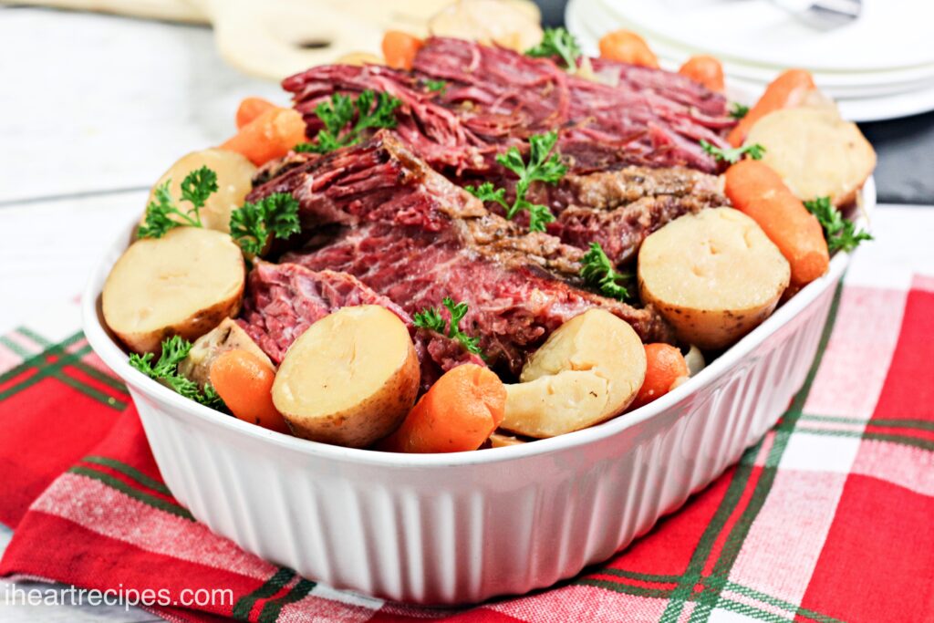 A large white casserole dish is filled with tender corned beef, surrounded by halved potatoes, carrots, and sprigs of fresh parsley.