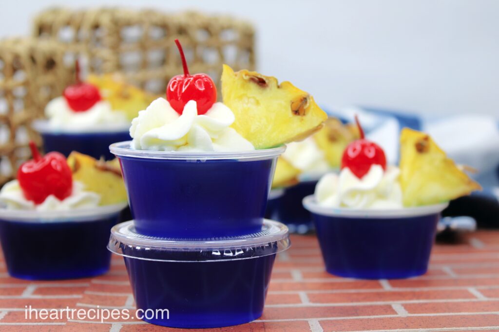 Small cups of Malibu Breeze Jello shots are stacked on a countertop. Each shot has blue Jello topped with a dollop of whipped cream, a cherry, and a pineapple wedge.