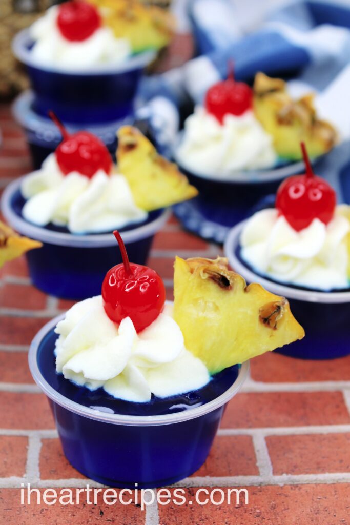 Cups of mini Malibu Breeze Jello shots sit on a brick surface. Each blue Jello shot is topped with whipped cream, pineapple, and a cherry.