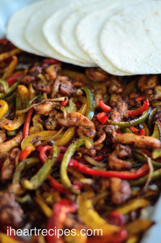 A close up image of a sheet pan filled with seasoned chicken fajitas with flour tortillas.