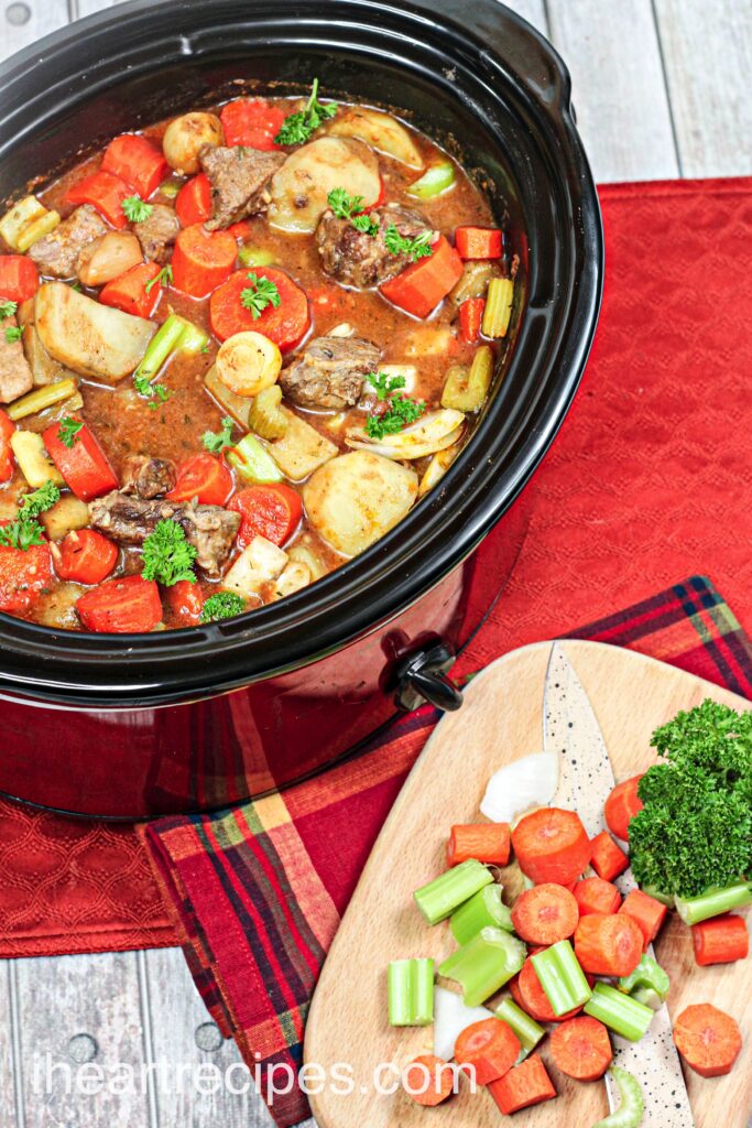 An overhead image of a large slow cooker filled with homemade beef stew sitting next to a cutting board filled with chopped celery and carrots.