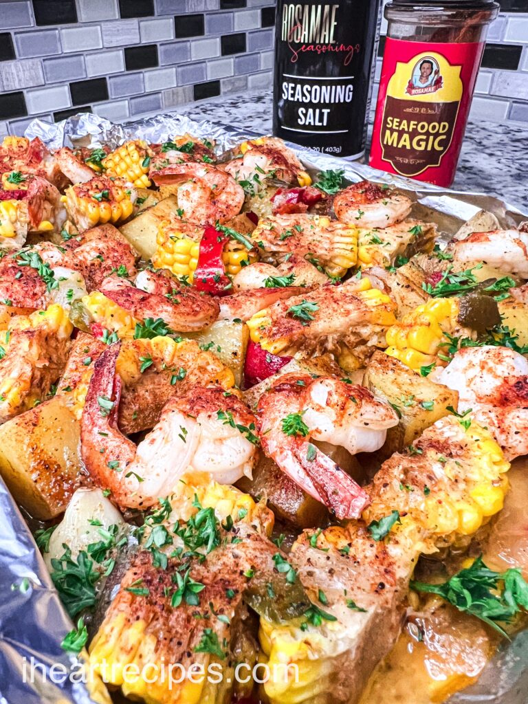 A sheet pan shrimp boil with vegetables and seasonings. Bottles of Rosamae Seafood Magic seasoning and seasoning salt sit on the counter next to the baking sheet.