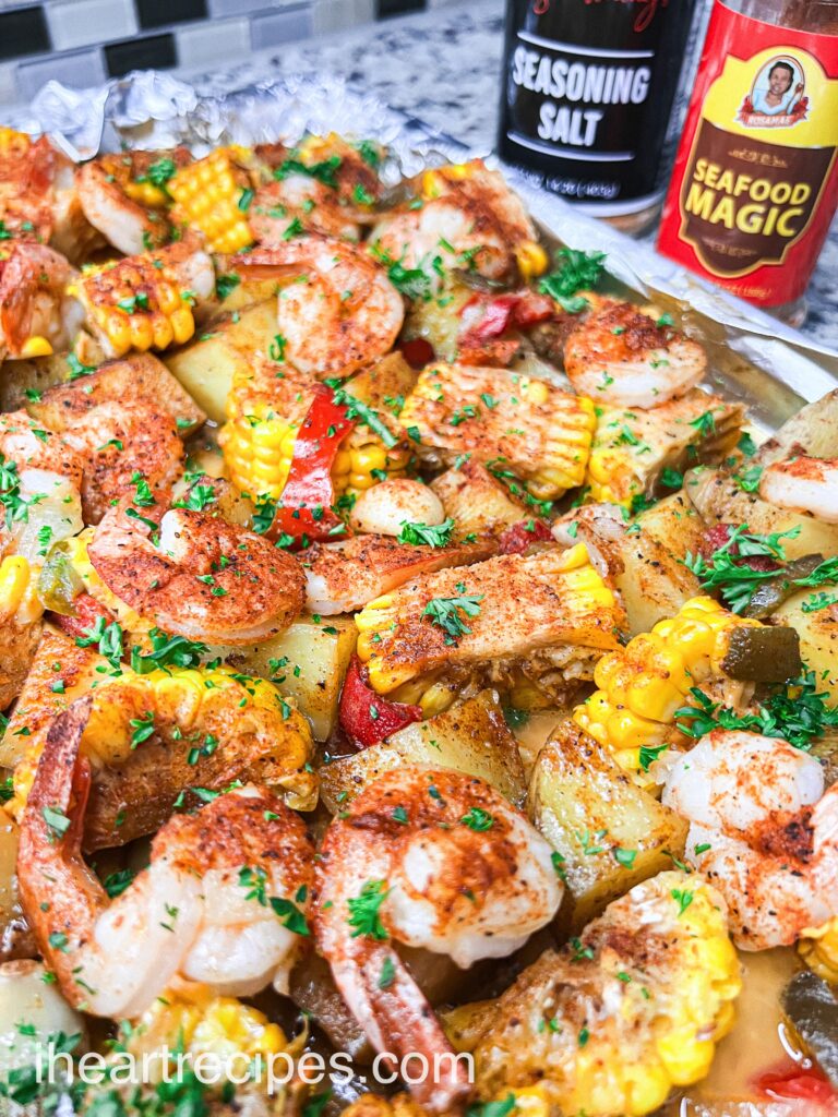 Ingredients for a shrimp sheet pan boil, spread out and seasoned on an aluminum foil lined pan. Shrimp, potatoes, corn, and peppers are covered with seafood seasoning and oil.