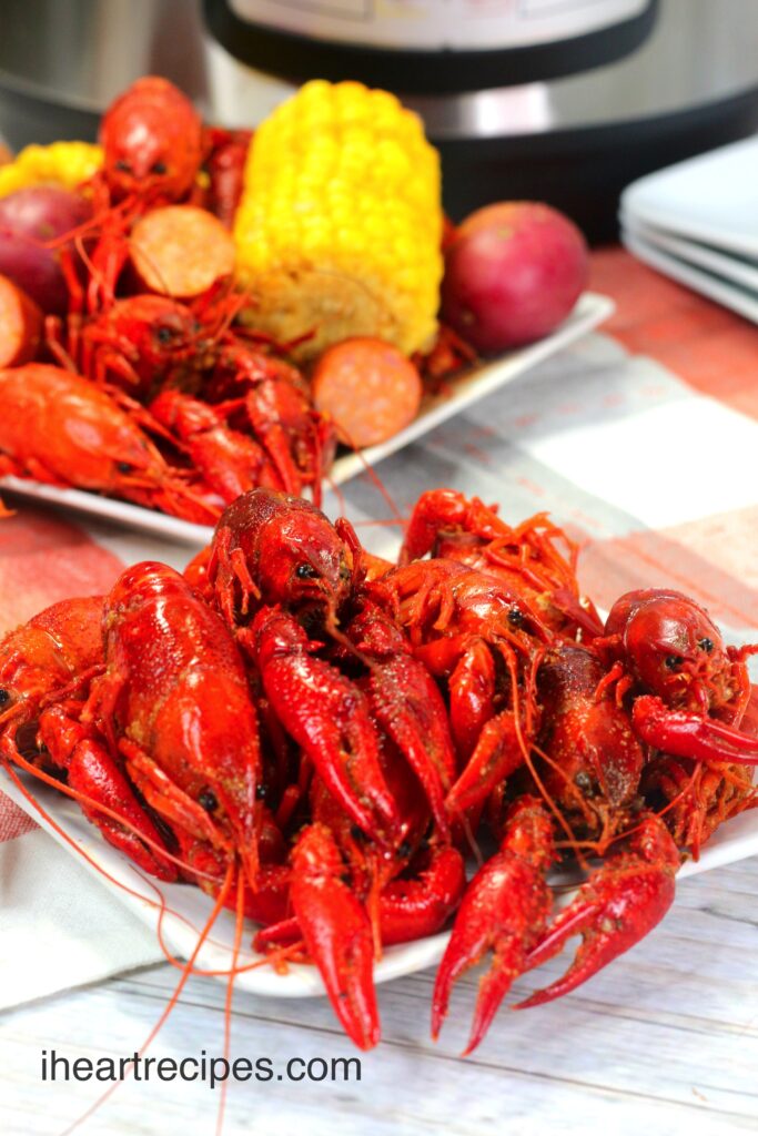 Two plates piled high with cooked crawfish. The plate in the background also has sausage, potatoes, and corn on the cob served along with the crawfish.