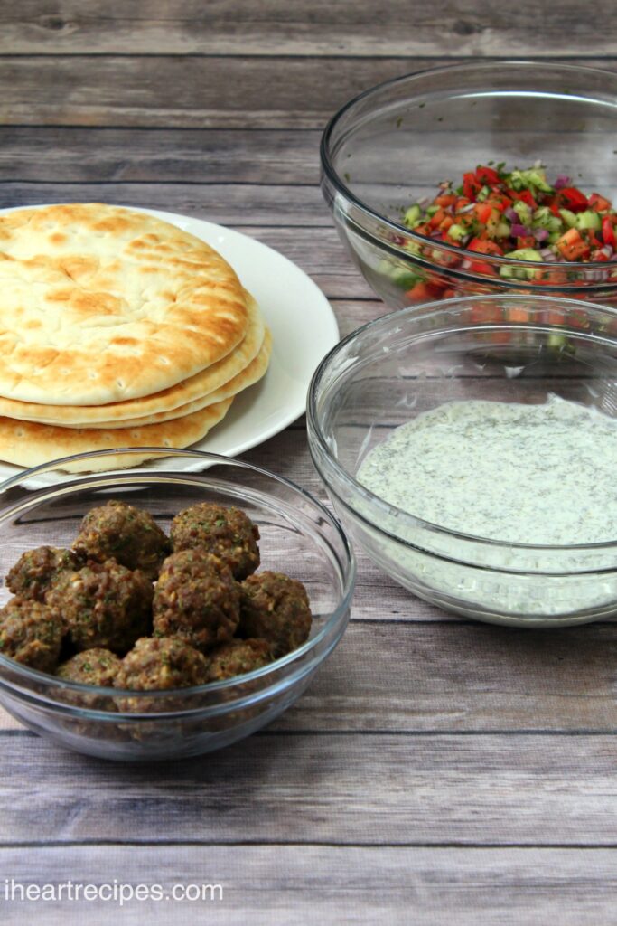 Recipe ingredients for Greek meatball gyros -- three bowls filled with meatballs, Cucumber Tomato Salad, and cucumber tomato salad, and a white plate of pita breads.