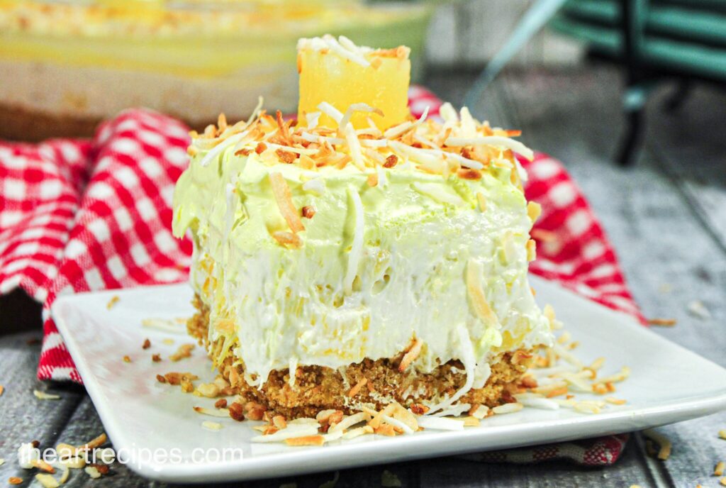 A slice of creamy no-bake pina colada cheesecake served on a white square plate. The cheesecake has a graham cracker crust and is topped with shredded toasted coconut and a pineapple square.