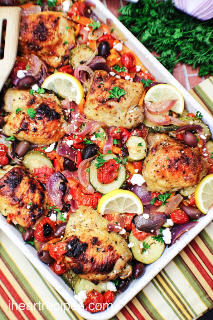 A close up overhead shot of Greek chicken baked on a sheet pan with a variety of vegetables, topped with Feta cheese and lemon slices.