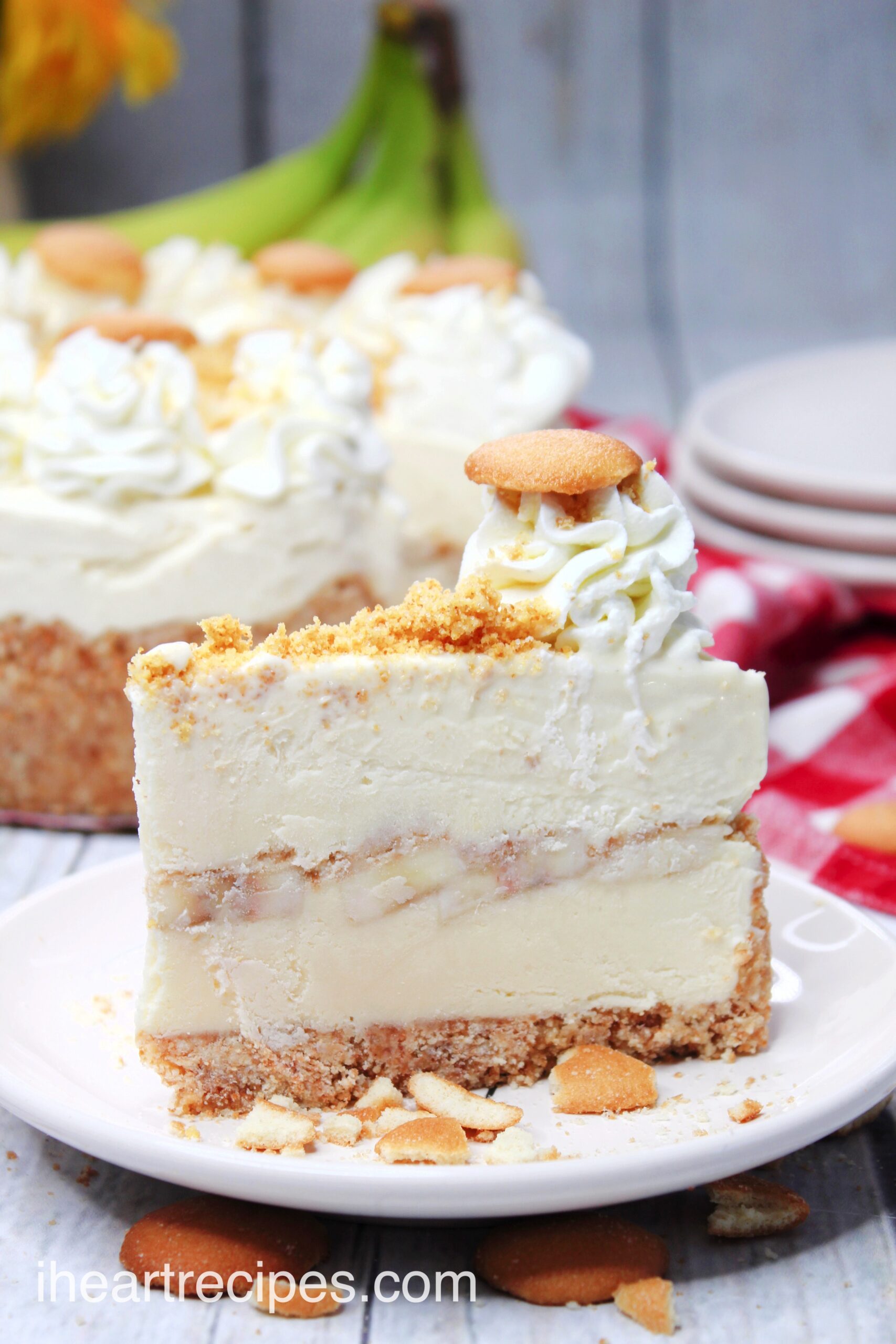 A single slice of banana pudding cheesecake. The slice has layers of ingredients - a vanilla wafer base, vanilla cheesecake, sliced bananas, and a banana pudding topping with whipped cream and crushed vanilla wafers.