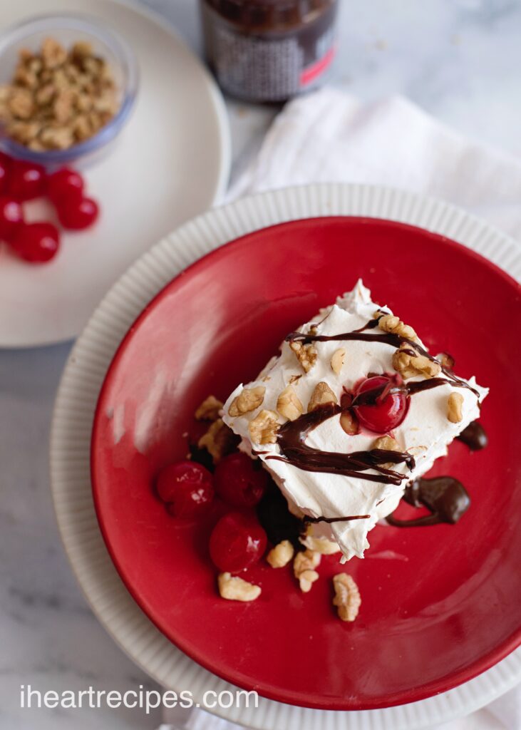 An overhead image of a slice of Banana Split Lasagna dessert, served on a red plate and topped with cherries, walnut pieces, and a fudge drizzle.