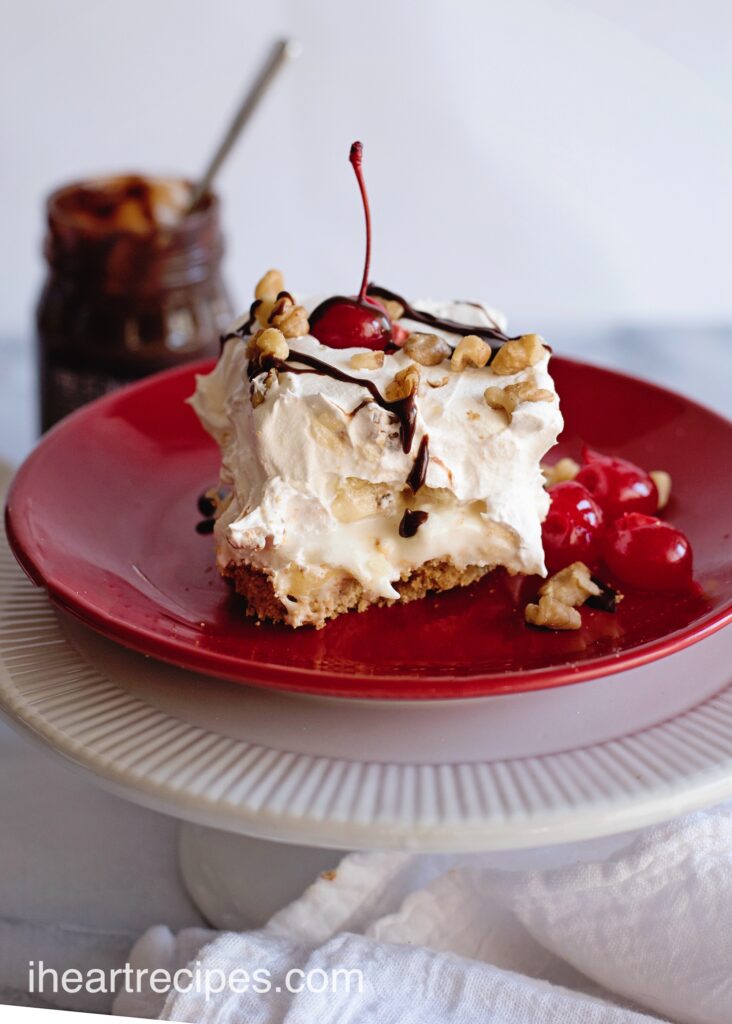 A slice of Banana Split Lasagna pictured from the side, showing layers of creamy banana filling, whipped topping, and a graham cracker base. The dessert is topped with walnuts, fudge drizzle, and cherries.